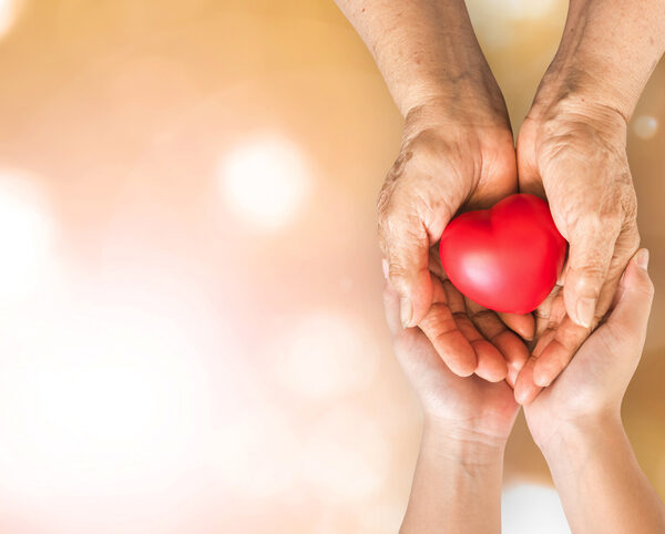 An elderly pair of hands holding a heart while a younger pair of hands supports from underneath