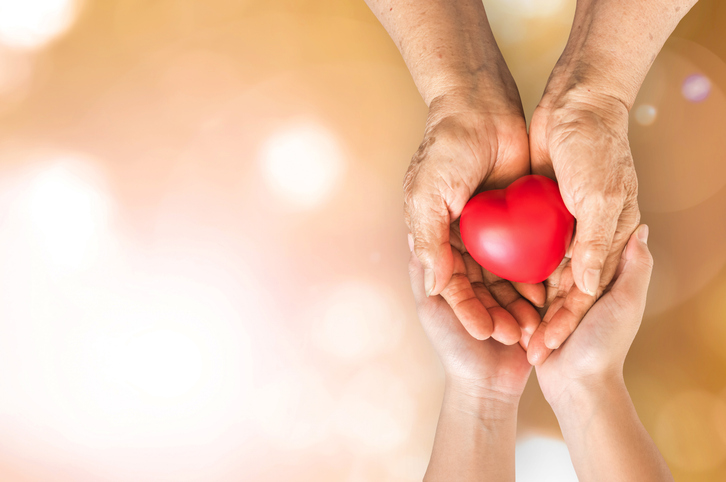 An elderly pair of hands holding a heart while a younger pair of hands supports from underneath