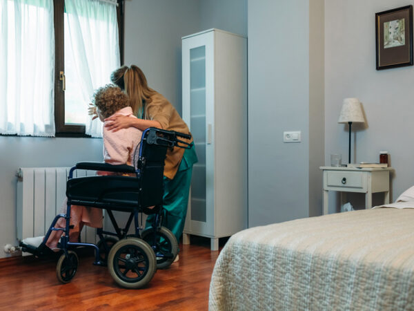 Female caregiver pulling back a curtain to show the outside to an older adult who is in a wheelchair