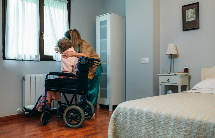 Female caregiver pulling back a curtain to show the outside to an older adult who is in a wheelchair