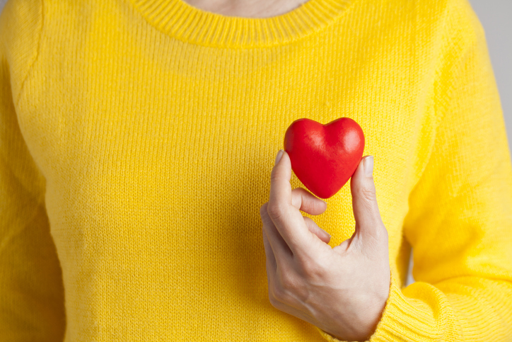 Woman wearing a bright yellow sweater holding a red heart up to her chest