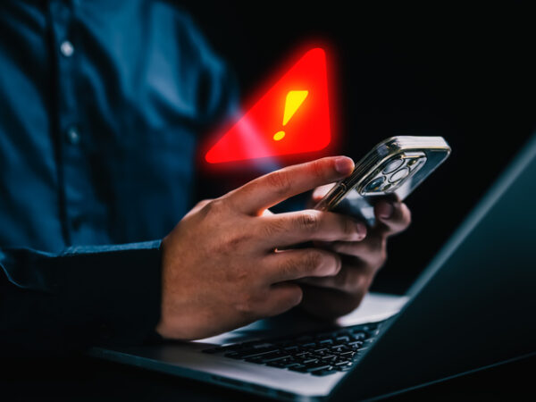 An older man in front of a laptop computer with his smartphone in his hand. A red warning exclamation point is hovering above his smartphone.