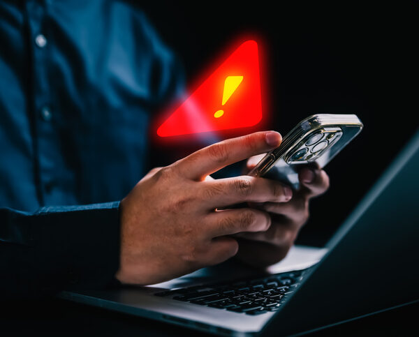 An older man in front of a laptop computer with his smartphone in his hand. A red warning exclamation point is hovering above his smartphone.