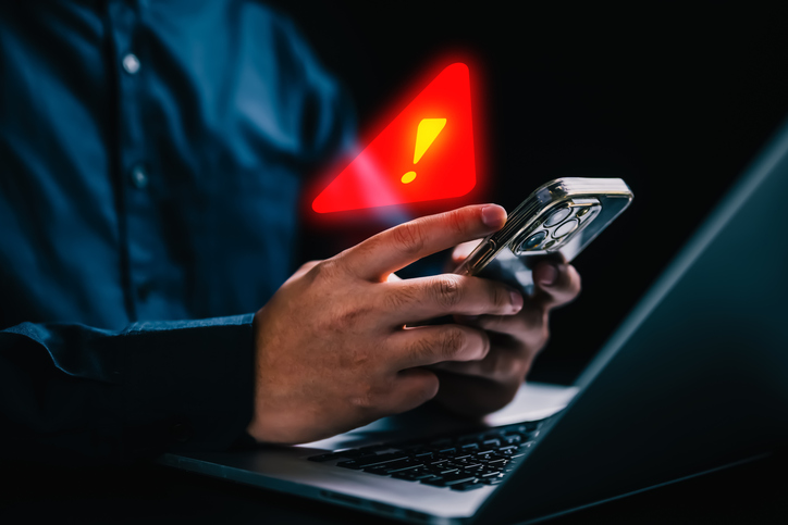 An older man in front of a laptop computer with his smartphone in his hand. A red warning exclamation point is hovering above his smartphone.