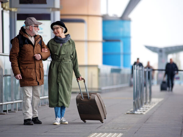 Older man and woman standing outside an airport. The woman is holding the handle of a suitcase.