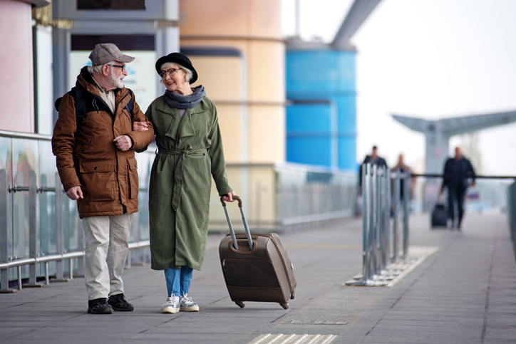 Older man and woman standing outside an airport. The woman is holding the handle of a suitcase.